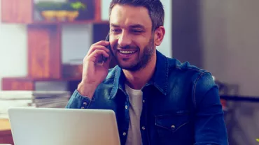 Hombre sonriendo mientras habla por teléfono y trabaja en laptop.