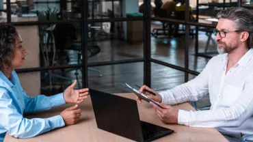 Dos personas en una reunión de trabajo, sentadas frente a frente en una oficina moderna.