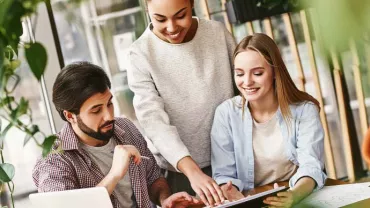 Tres personas colaboran en un proyecto en una mesa, una sobre una laptop.