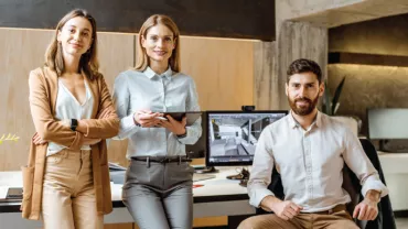 Tres personas en una oficina moderna, sonrientes y de pie junto a un escritorio con computadora.