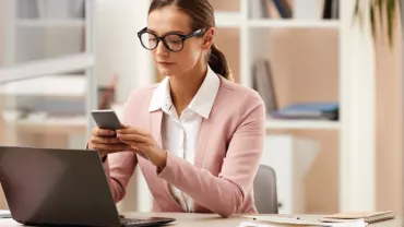 Mujer en oficina usando teléfono frente a computadora portátil.