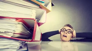 Persona sorprendida mirando una pila de carpetas llenas de papeles.