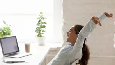 Mujer estirándose frente a una laptop en una oficina luminosa.
