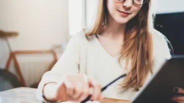 Mujer con gafas usando una tableta mientras toma notas en un escritorio.