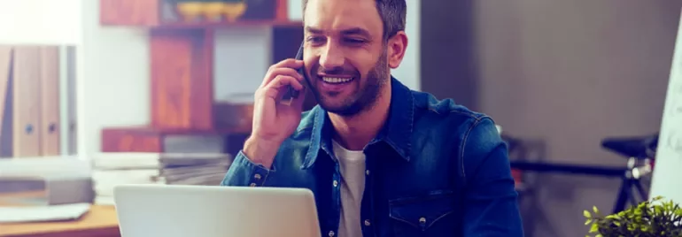 Hombre sonriendo mientras habla por teléfono y trabaja en laptop.