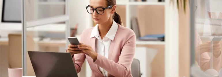 Mujer en oficina usando teléfono frente a computadora portátil.