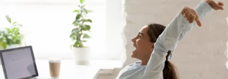 Mujer estirándose frente a una laptop en una oficina luminosa.