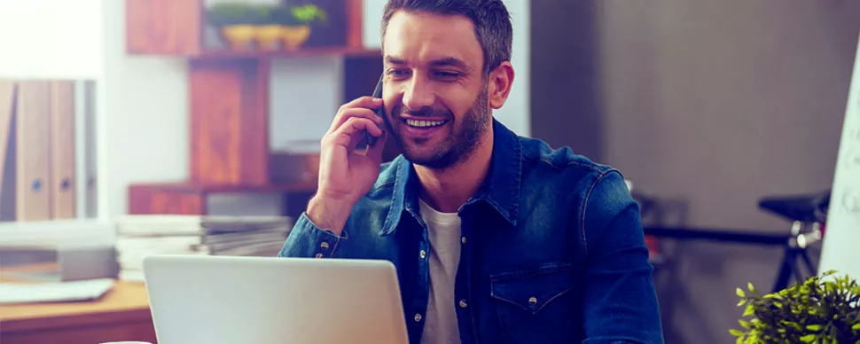Hombre sonriendo mientras habla por teléfono y trabaja en laptop.