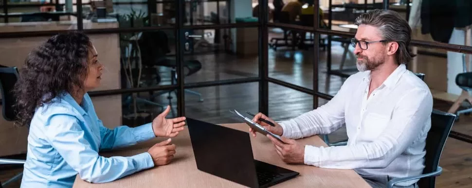 Dos personas en una reunión de trabajo, sentadas frente a frente en una oficina moderna.