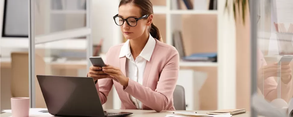 Mujer en oficina usando teléfono frente a computadora portátil.