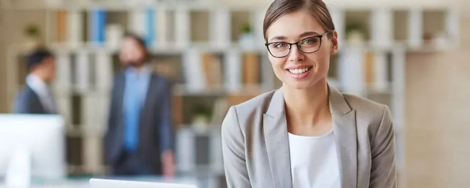 Mujer sonriente con gafas y traje gris en una oficina.