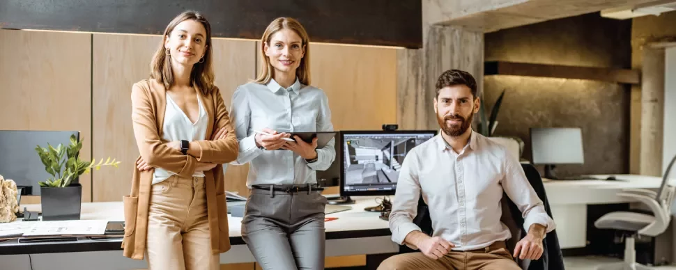 Tres personas en una oficina moderna, sonrientes y de pie junto a un escritorio con computadora.