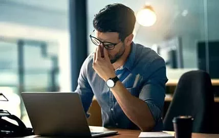 Hombre con gafas frente a laptop, parece cansado.