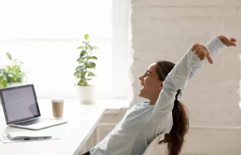Mujer estirándose frente a una laptop en una oficina luminosa.