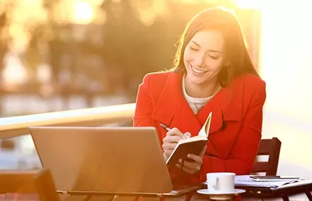 Mujer sonríe mientras toma notas frente a una laptop al atardecer.