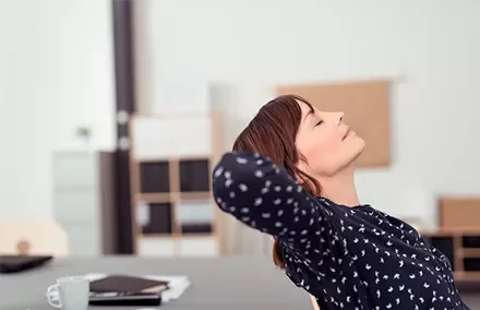 Mujer relajándose en una oficina, con los ojos cerrados y manos detrás de la cabeza.